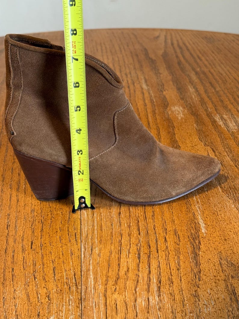 May include: A brown suede ankle boot with a pointed toe and a stacked heel. The boot is approximately 8 inches tall, measured with a yellow tape measure. The boot has a V-shaped topline and a textured panel on the back.