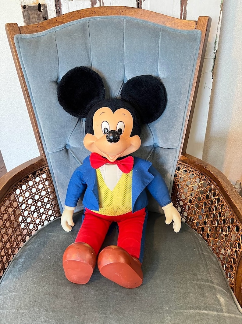 K&ouml;nnte beinhalten: Eine Pl&uuml;sch-Mickey-Mouse-Puppe, die einen blauen Anzug mit einer gelben Weste und roten Hosen tr&auml;gt, sitzt auf einem grauen Stuhl mit einem Holzrahmen.