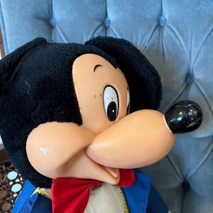 K&ouml;nnte beinhalten: Eine Vintage-Pl&uuml;schpuppe von Mickey Mouse, die einen blauen Anzug mit einer roten Schleife und einem wei&szlig;en Kragen tr&auml;gt. Die Puppe hat schwarzes Fell, eine braune Nase und gro&szlig;e schwarze Augen.
