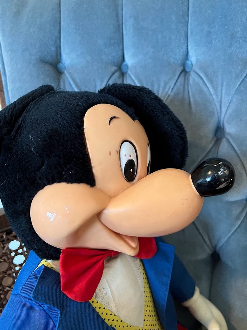 K&ouml;nnte beinhalten: Eine Vintage-Pl&uuml;schpuppe von Mickey Mouse, die einen blauen Anzug mit einer roten Schleife und einem wei&szlig;en Kragen tr&auml;gt. Die Puppe hat schwarzes Fell, eine braune Nase und gro&szlig;e schwarze Augen.