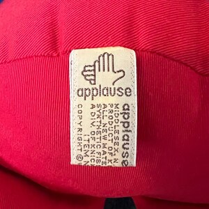 K&ouml;nnte beinhalten: Ein rotes und blaues Pl&uuml;schtier mit einem wei&szlig;en Etikett, auf dem "applause" steht und der Text "COPYRIGHT 1987 ALL RIGHTS RESERVED" und "A DIV. OF KNICK-KNACK, INC. ITEM NO. 1234567890" enthalten ist.