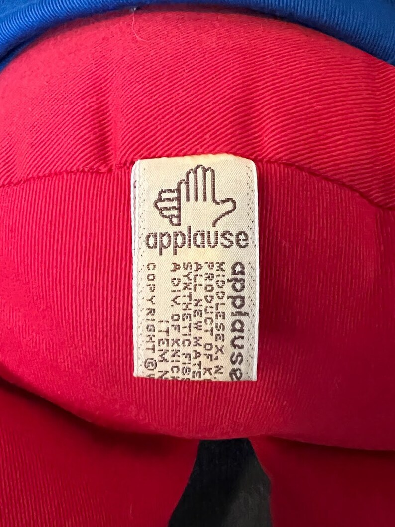 K&ouml;nnte beinhalten: Ein rotes und blaues Pl&uuml;schtier mit einem wei&szlig;en Etikett, auf dem "applause" steht und der Text "COPYRIGHT 1987 ALL RIGHTS RESERVED" und "A DIV. OF KNICK-KNACK, INC. ITEM NO. 1234567890" enthalten ist.