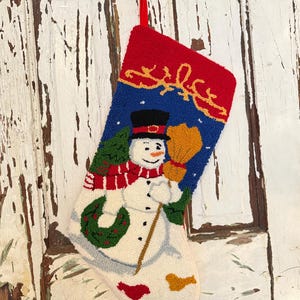 Puede incluir: Un calcetín navideño con un diseño de muñeco de nieve. El calcetín presenta una parte superior roja, un centro azul con un muñeco de nieve sosteniendo una escoba y una base blanca. El muñeco de nieve lleva un sombrero negro, una bufanda roja y una corona verde.