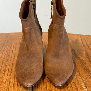 May include: A pair of brown suede ankle boots with pointed toes. The boots have a side zipper and a stacked heel. The boots are displayed on a wooden surface.