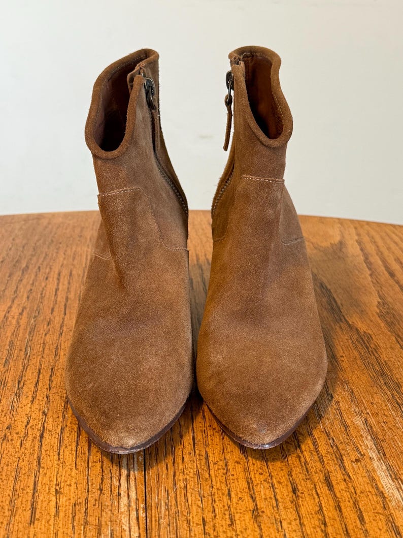 May include: A pair of brown suede ankle boots with pointed toes. The boots have a side zipper and a stacked heel. The boots are displayed on a wooden surface.
