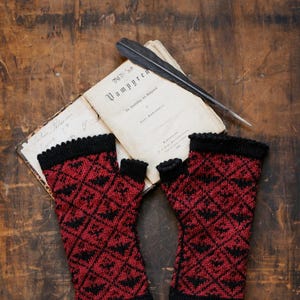 May include: A pair of red and black fingerless gloves with a bat pattern. The gloves are lying on a wooden surface next to an open book with the title "Vampyren" on the cover.