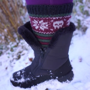 May include: Black winter boot with a zipper, faux fur trim, and a patterned sock peeking out. The sock features a snowflake design in white and burgundy, with green and burgundy stripes. The boot is covered in snow.