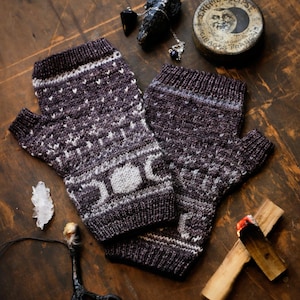 Könnte beinhalten: Ein Paar braune und weiße gestrickte Fingerlose Handschuhe mit einem Mond- und Sternenmuster. Die Handschuhe liegen auf einer Holzoberfläche mit anderen Gegenständen, darunter ein Kristall, ein Vogelbein und ein kleines Holzkreuz.