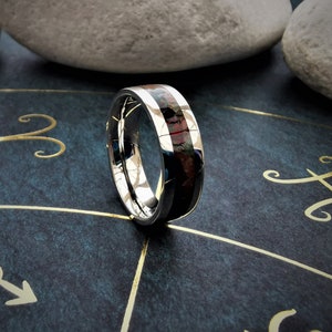 May include: A silver ring with a black and red inlay. The ring is on a dark blue surface with gold zodiac symbols.