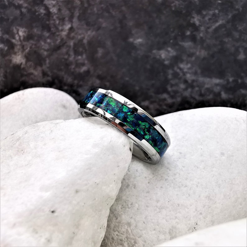 May include: A silver ring with a green and blue opal inlay. The ring is set on a white rock.