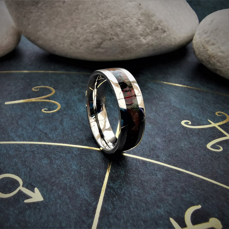 May include: A silver ring with a black and red inlay. The ring is on a dark blue surface with gold zodiac symbols.