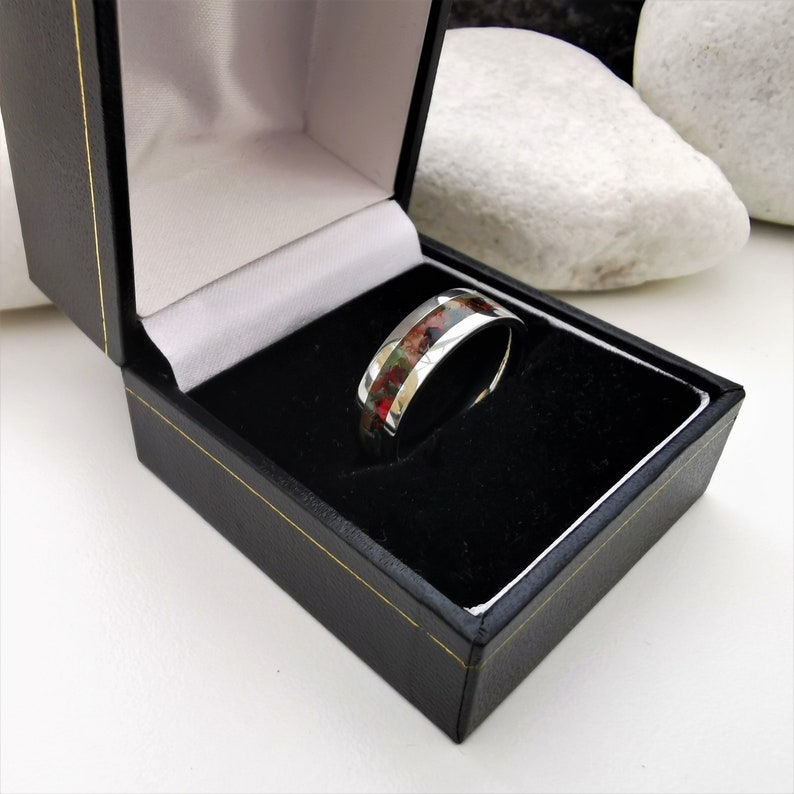 May include: A silver ring with a thin band of red, green, and white inlay. The ring is in a black ring box.