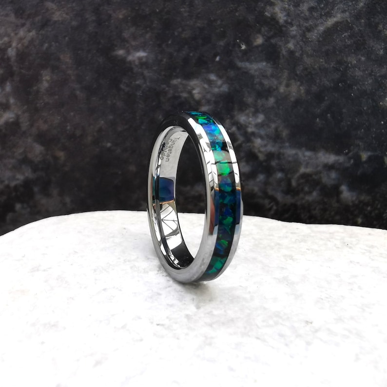 May include: A silver tungsten carbide ring with a thin band of green and blue opal inlay. The ring is engraved with "Tungsten Carbide" on the inside of the band.