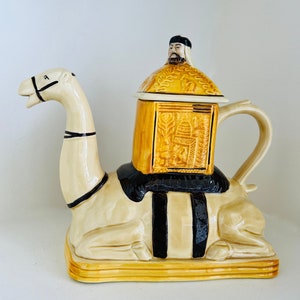 May include: A ceramic teapot shaped like a camel with a detailed, ornate lid featuring a man's face. The teapot is decorated with black and gold accents and has a spout and handle.