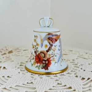 May include: A white porcelain bell featuring a hand-painted design of a colorful bird perched on a branch with orange and yellow flowers. The bell has a gold-trimmed base and a decorative handle at the top, showcasing intricate detail and craftsmanship.