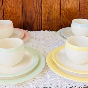 May include: Four sets of pastel-colored teacups and saucers. Each cup has a colored band around the top and a matching handle. The saucers are also color-coordinated. The colors include pink, green, blue, and yellow.