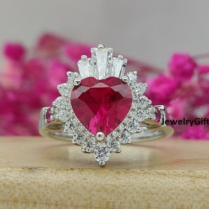 May include: A silver ring with a large heart-shaped ruby surrounded by smaller diamonds. The ring has a delicate, intricate design with a crown-like top.