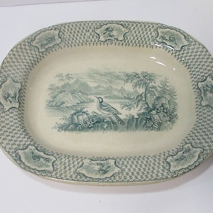 Puede incluir: Un plato de servir ovalado vintage con un diseño verde y blanco. El centro presenta un pájaro en una escena de paisaje. El borde tiene un patrón repetitivo de hojas verdes y un patrón de diamante.