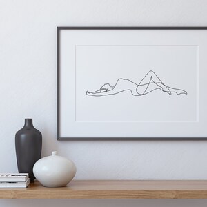 May include: A black and white line drawing of a person lying down, framed in a black frame and displayed on a shelf with a black vase and a white vase.