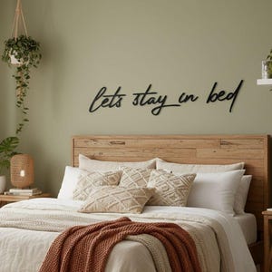 Op de afbeelding: Een zwart metalen wandbord met de cursieve tekst "lets stay in bed" boven een houten hoofdeinde. Het bed is opgemaakt met crèmekleurig beddengoed, een textuurrijke roestkleurige sprei en diverse decoratieve kussens in neutrale tinten.