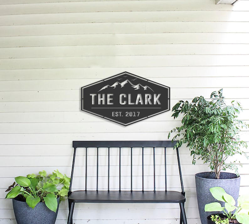 Op de afbeelding: Een donkergrijs metalen bord met de tekst "THE CLARK" en "EST. 2017" in een bergthema. Het bord is bevestigd aan een witte houten muur boven een zwarte bank en potplanten.