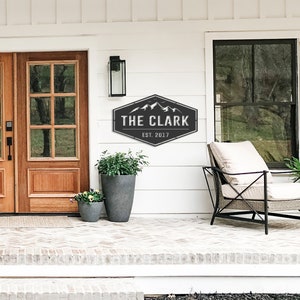 Op de afbeelding: Een zwart-wit metalen bord met de tekst "THE CLARK EST. 2017" en een bergketenafbeelding. Het bord is bevestigd aan een witte muur boven een bakstenen veranda, naast een houten deur en een plant in een pot.