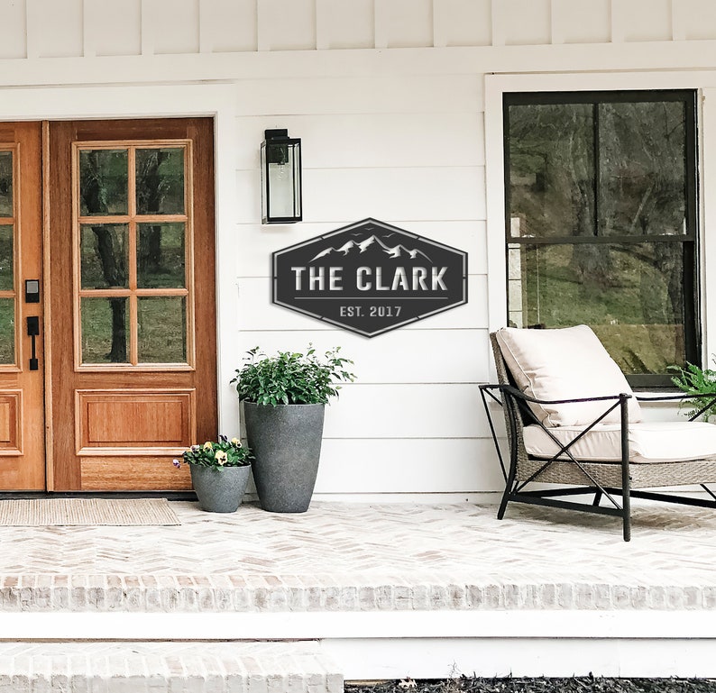 Op de afbeelding: Een zwart-wit metalen bord met de tekst "THE CLARK EST. 2017" en een bergketenafbeelding. Het bord is bevestigd aan een witte muur boven een bakstenen veranda, naast een houten deur en een plant in een pot.