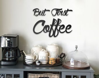 Może przedstawiać: Czarny metalowy znak z napisem "But First Coffee" wiszący na białej ścianie nad szarą szafką z ekspresem do kawy, pojemnikami i szklaną kopułą na górze.