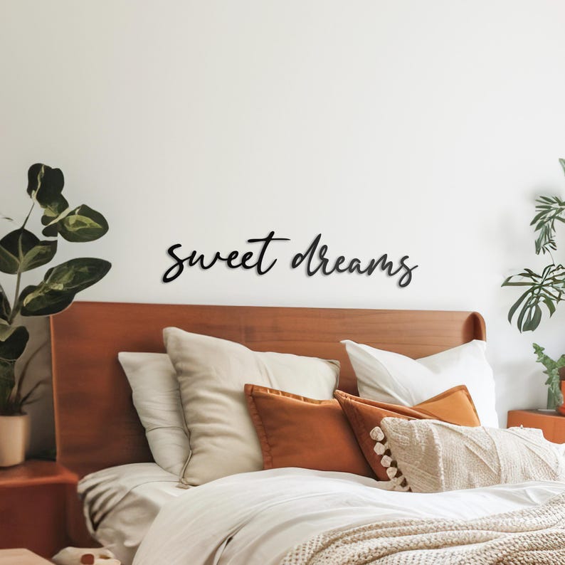May include: Black wooden wall decor with the words "sweet dreams" in a cursive font. The decor is above a bed with white bedding and brown throw pillows.