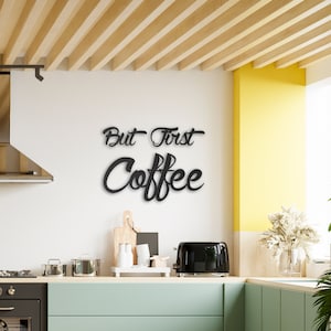 May include: Black metal wall decor with the text "But First Coffee" in a stylized font. The decor is mounted on a white wall in a kitchen setting.