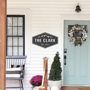 Op de afbeelding: Een zwart en grijs metalen bord met de tekst "THE CLARK" en "EST. 2017" is op een witte muur gemonteerd. Het bord heeft een bergketenontwerp. Een veranda met een zwarte bank, een deur en een krans is zichtbaar.