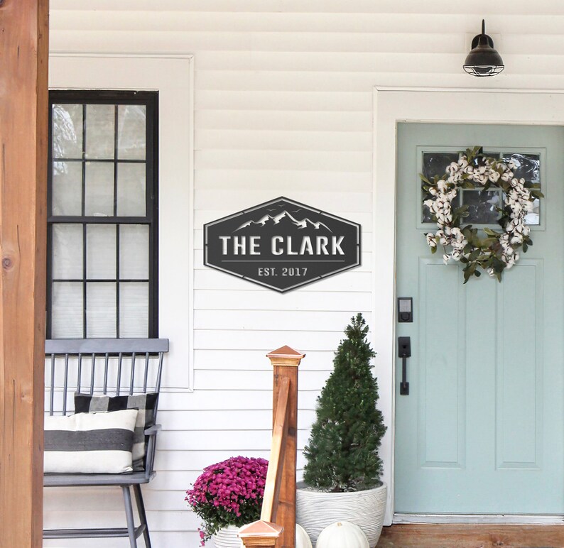 Op de afbeelding: Een zwart en grijs metalen bord met de tekst "THE CLARK" en "EST. 2017" is op een witte muur gemonteerd. Het bord heeft een bergketenontwerp. Een veranda met een zwarte bank, een deur en een krans is zichtbaar.