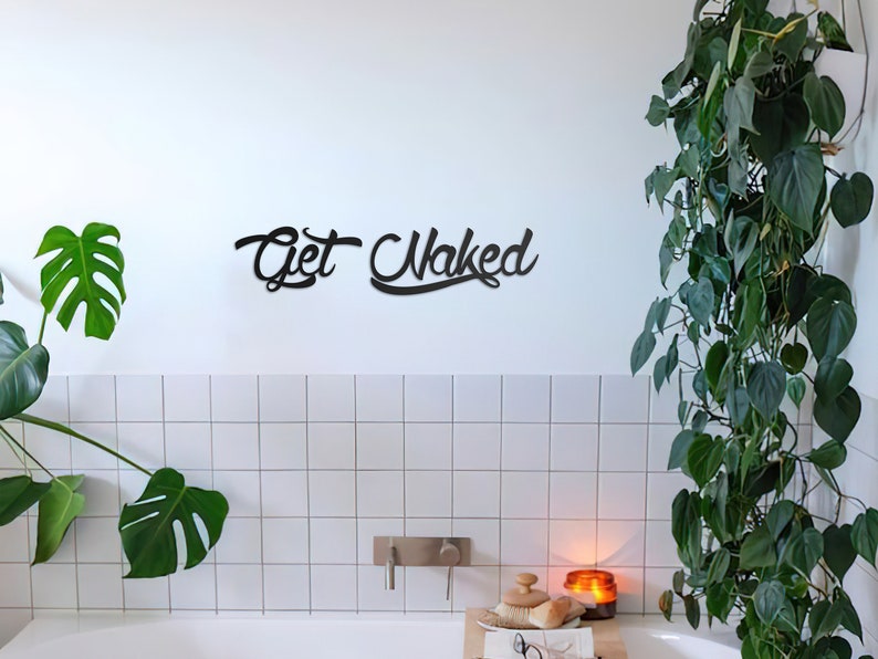 May include: A black metal sign with the text "Get Naked" in a cursive font, hanging on a white tiled wall in a bathroom. The sign is above a white bathtub and a white countertop with a candle, a brush, and glasses on it.