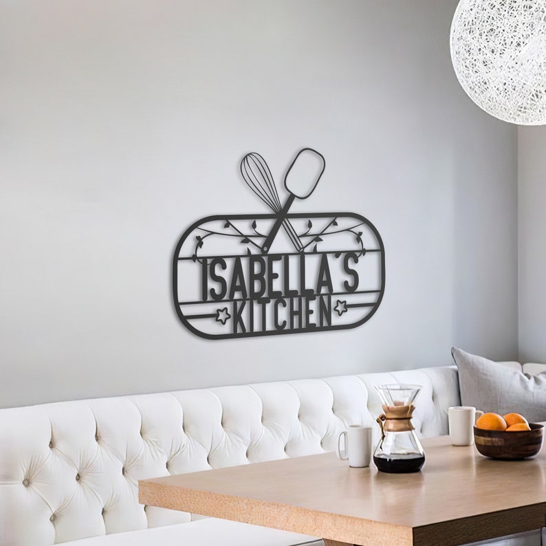 May include: Black metal kitchen sign with the text "Isabella's Kitchen" and crossed whisk and spatula. The sign is mounted on a white wall above a white couch and a wooden table.