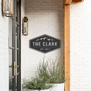 Op de afbeelding: Een zwart metalen bord met de tekst "THE CLARK" en "EST. 2017" in een zeshoekige vorm. Het heeft een berglandschap. Het bord is bevestigd aan een witte bakstenen muur. Buitenverlichting en potplanten zijn ook zichtbaar.