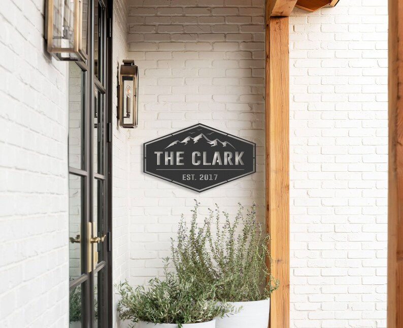Op de afbeelding: Een zwart metalen bord met de tekst "THE CLARK" en "EST. 2017" in een zeshoekige vorm. Het heeft een berglandschap. Het bord is bevestigd aan een witte bakstenen muur. Buitenverlichting en potplanten zijn ook zichtbaar.