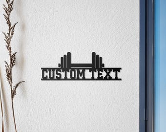 May include: Black metal wall decor featuring a barbell with weights on each end. The barbell has a rectangular sign below it that says "CUSTOM TEXT".