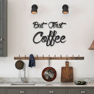 May include: Black metal wall decor with the text "But First Coffee" in a cursive font. The decor is hanging on a white wall in a kitchen setting.