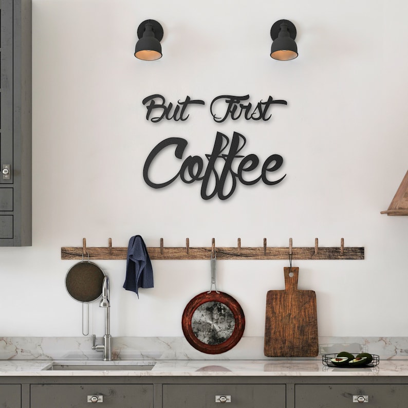 May include: Black metal wall decor with the text "But First Coffee" in a cursive font. The decor is hanging on a white wall in a kitchen setting.