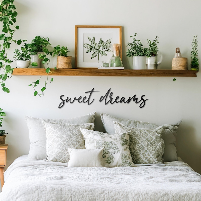 May include: A white bed with a white duvet cover and several patterned throw pillows. The bed is against a white wall with a wooden shelf above it. The shelf is decorated with plants and a framed print. The words "sweet dreams" are written in black script on the wall above the bed.
