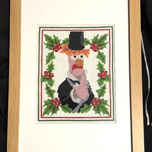 May include: Cross-stitch artwork of a turkey wearing a top hat and tuxedo, framed in a light wood frame. The turkey is surrounded by a wreath of green leaves and red berries, with a red and green border.