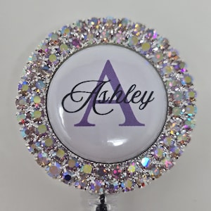 May include: A round badge reel with a white center featuring the name "Ashley" in cursive and a purple letter "A". The badge is surrounded by a ring of iridescent rhinestones.