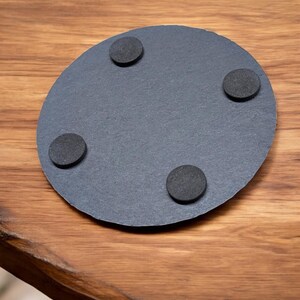 以下が含まれることがあります： 黒色のスレート製の丸いコースターで、底面に黒いゴム製の足が4つ付いている