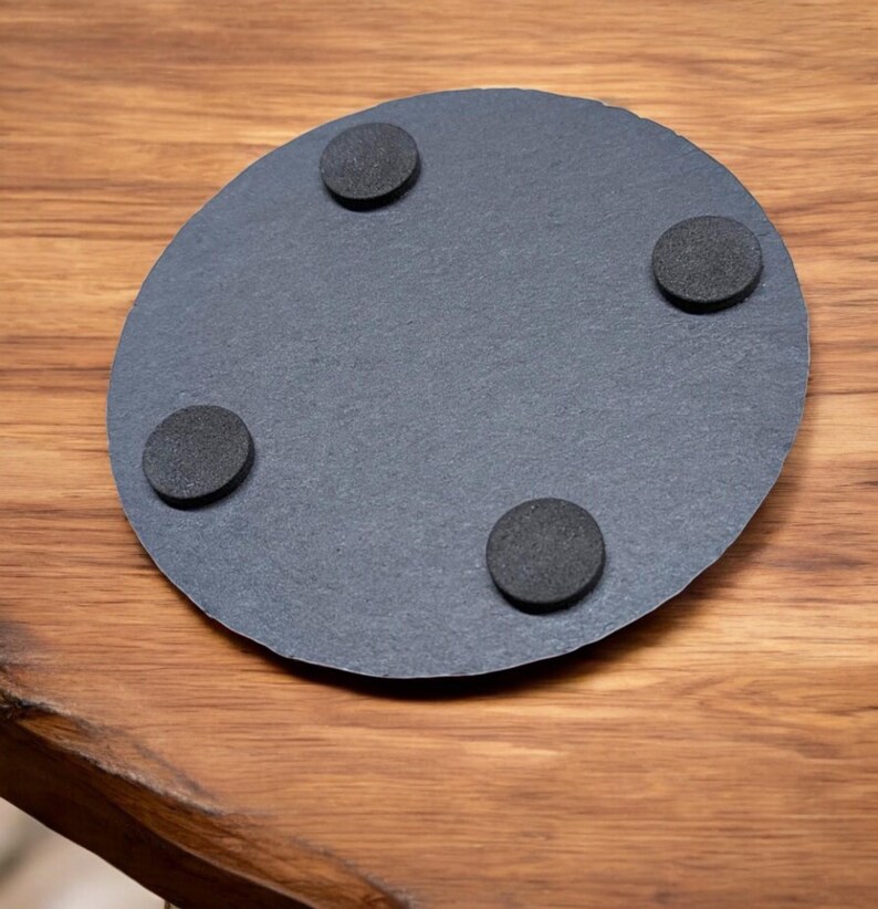 以下が含まれることがあります： 黒色のスレート製の丸いコースターで、底面に黒いゴム製の足が4つ付いている