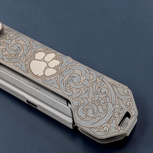 May include: Close-up of a multi-tool with a decorative design. The tool features a paw print and ornate scrollwork in shades of gray and silver. The design is etched into the metal surface, creating a detailed and textured appearance.