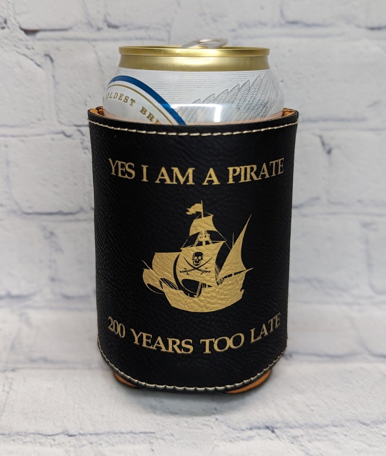 Pu&ograve; includere: Frigorifero per lattine in pelle nera con testo dorato e un'immagine di una nave pirata dorata. Il testo recita "S&igrave;, sono un pirata, 200 anni troppo tardi".