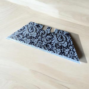May include: A silver utility knife blade with a black and silver floral pattern.