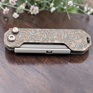 May include: A small, silver and bronze multi-tool with an ornate floral pattern engraved on the top. The tool has a rectangular shape with rounded edges and visible screws. The design features roses and swirling leaves.
