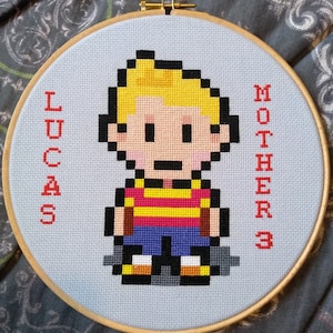 May include: Cross-stitch embroidery of a pixelated character named Lucas from the video game EarthBound, also known as Mother 3. The embroidery is framed in a wooden hoop and has the words "Lucas" and "Mother 3" stitched in red on the left and right sides.