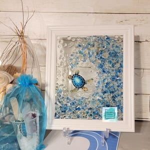 May include: A framed glass art piece featuring a blue and yellow sea turtle swimming in a wave of blue and clear glass beads. The frame is white and the glass beads are arranged in a wave shape.